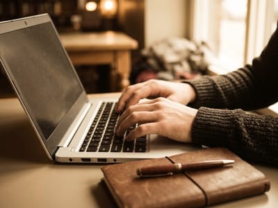 Hands typing on a laptop next to a leather journal and pen in a warm cafe