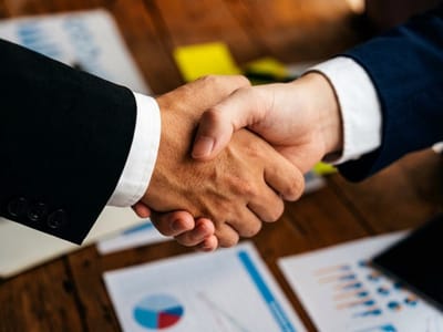 Two people in suits shaking hands above a table of printed charts and sticky notes