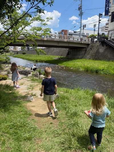Three children walking along a grassy riverbank in a Japanese town with a bridge in the background