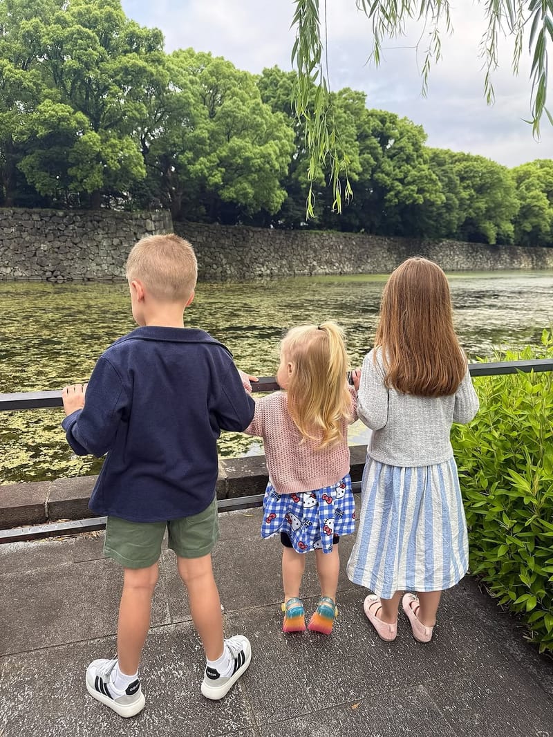Three children leaning on a railing overlooking a pond with weeping willows and a stone wall