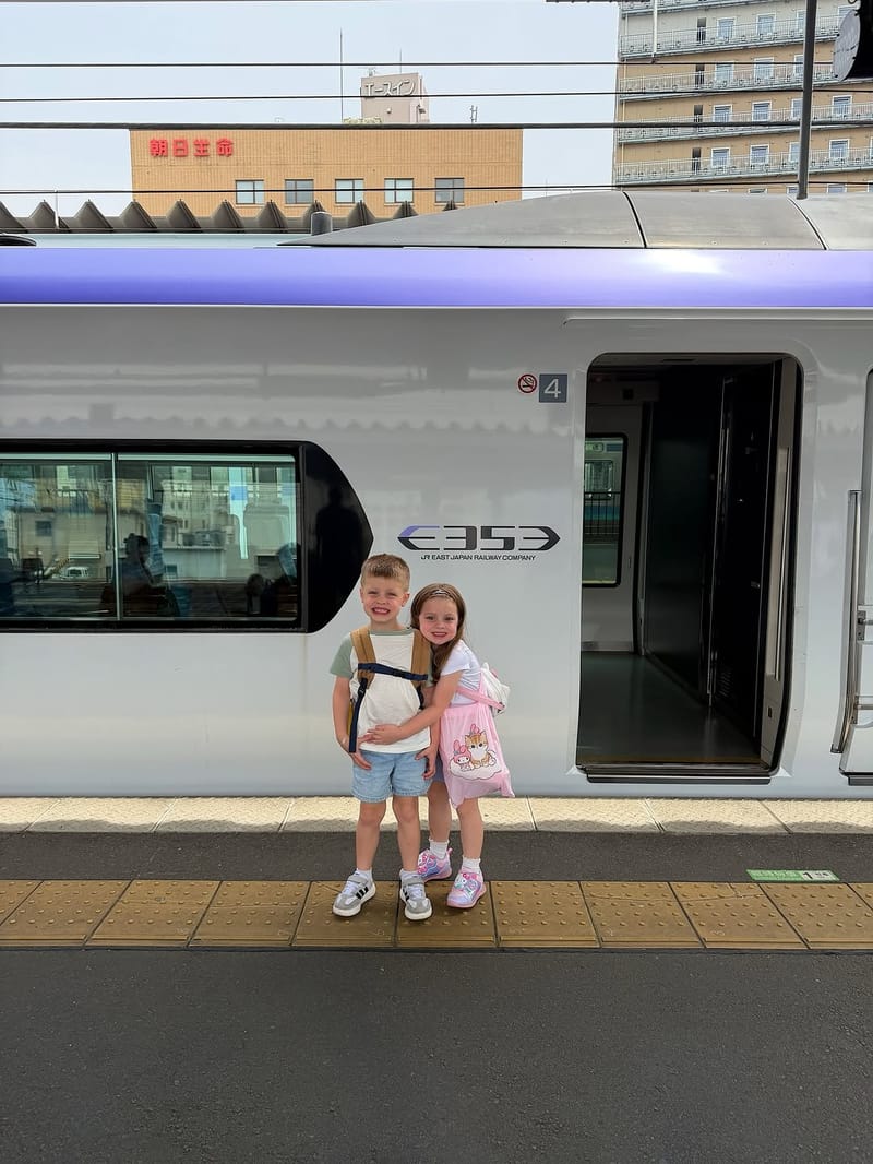 Two children smiling and posing in front of a sleek JR East train at a Japanese platform